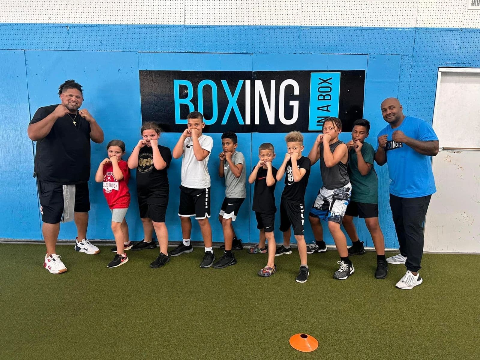 Nine kids in a fists-up line with two coaches under the Boxing In A Box sign